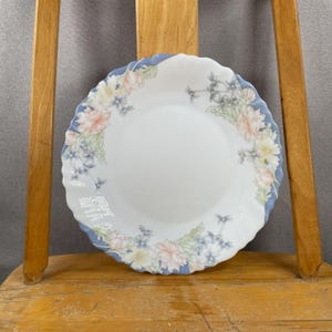 May include: White ceramic plate with a floral design. The plate has a wavy edge with a light blue border. The floral pattern includes pink, yellow, and blue flowers with green leaves.