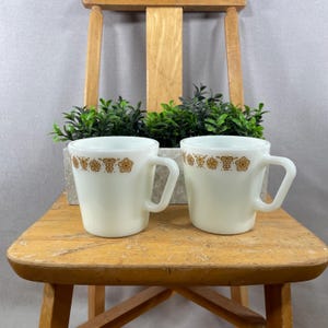 May include: Two white ceramic mugs with a brown floral pattern around the rim. The mugs have a simple, curved handle and are set on a wooden stool. A small planter with green plants is behind the mugs.
