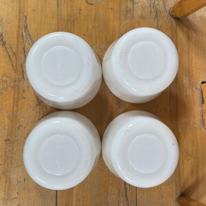 May include: Four white, round, ceramic bowls are arranged on a wooden surface. The bowls have a simple, classic design with a slightly raised center and a smooth, glossy finish. The bowls are likely for serving or eating.