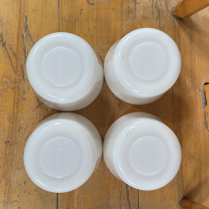 May include: Four white, round, ceramic bowls are arranged on a wooden surface. The bowls have a simple, classic design with a slightly raised center and a smooth, glossy finish. The bowls are likely for serving or eating.