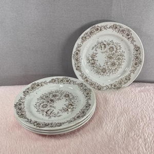 May include: Four white ceramic plates with a floral design. Each plate has a brown floral wreath around the edge and a central floral arrangement. The plates have a speckled texture.