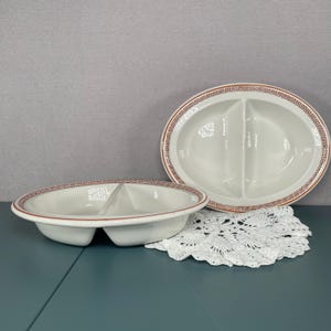 May include: Two white oval divided serving dishes with a red and brown Greek key border. One dish is in the foreground, and the other is in the background. The dishes are on a white lace doily.
