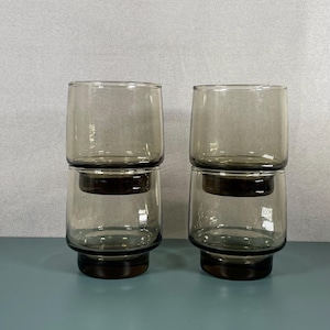 May include: Four stacked, smoky gray drinking glasses. Each glass has a cylindrical top and a dark brown base. The glasses are arranged in two pairs, with one glass stacked on top of another. The background is a light gray.