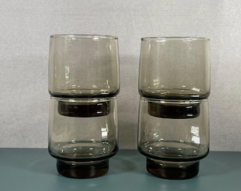 May include: Four stacked, smoky gray drinking glasses. Each glass has a cylindrical top and a dark brown base. The glasses are arranged in two pairs, with one glass stacked on top of another. The background is a light gray.