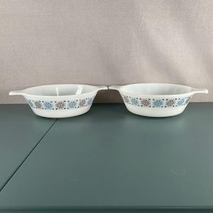 May include: Two white glass baking dishes with handles, each featuring a repeating pattern of blue and black geometric snowflake-like designs around the exterior rim. The bowls have a smooth, rounded shape and a glossy finish.