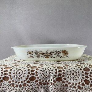 Puede incluir: Una fuente ovalada blanca con un diseño floral en marrón y dorado. La fuente descansa sobre un mantel de encaje blanco. El patrón floral incluye flores y hojas estilizadas.