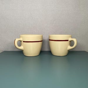 Op de afbeelding: Twee crèmekleurige keramische mokken met een donkerrode horizontale streep. De mokken hebben afgeronde lichamen en een gebogen handvat. De mokken staan op een turquoise oppervlak.
