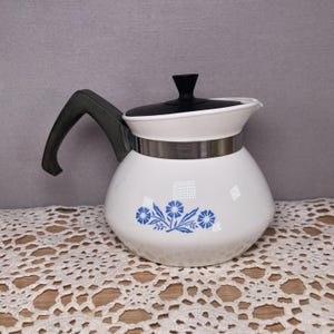 May include: White ceramic teapot with a black handle and lid. The teapot features a silver band and a blue floral design. The teapot is sitting on a white lace doily.