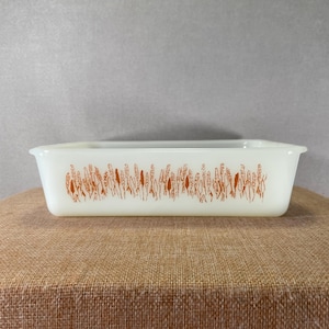 Könnte beinhalten: Rechteckige, cremefarbene Pyrex-Backform mit einem weizenähnlichen Muster in Orange. Die Form hat sanft geschwungene Seiten und ein schlichtes, funktionales Design. Das Weizenmuster wiederholt sich an der Seite der Form.
