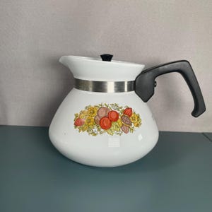 May include: White ceramic coffee pot with a black handle and lid. The pot features a decorative silver metal band and a colourful floral design with fruits and flowers. The pot has a spout for pouring.