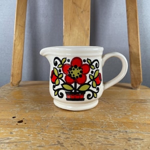 Könnte beinhalten: Ein cremefarbenes Milchkännchen aus Keramik mit einem zentralen roten Blumenmotiv, einem grünen Blütenkern, schwarzen Stielen und grünen Blättern. Das Kännchen hat einen bauchigen Körper, einen kleinen Ausguss und einen geschwungenen Henkel.
