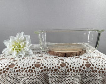 May include: A clear glass rectangular baking dish with handles, resting on a wooden slice. A white flower is placed to the left of the dish. The dish sits on a white lace tablecloth.