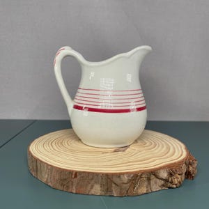 May include: A small, white ceramic pitcher with a handle and a spout. The pitcher is decorated with horizontal red stripes around the body. It sits on a round, natural wood slice.