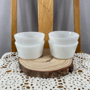 May include: Four white, stackable, small bowls. The bowls are made of a solid white material and have a rounded top and a slightly flared base. They are arranged in two stacks of two on a wooden slice.