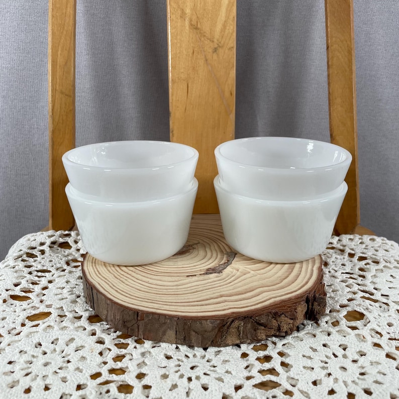 May include: Four white, stackable, small bowls. The bowls are made of a solid white material and have a rounded top and a slightly flared base. They are arranged in two stacks of two on a wooden slice.