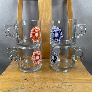 May include: Four clear glass mugs with handles are stacked. Two mugs have an orange and white floral design, and two have a blue and white floral design. The mugs are on a wooden surface.