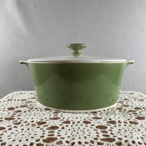 May include: A green ceramic casserole dish with a white lid. The dish has a rounded shape with two small handles and a knob on the lid. The dish is sitting on a white lace tablecloth.