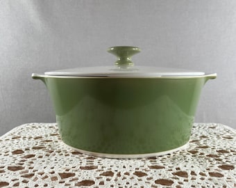 May include: A green ceramic casserole dish with a white lid. The dish has a rounded shape with two small handles and a knob on the lid. The dish is sitting on a white lace tablecloth.