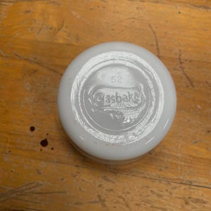 May include: A white, round Glasbake lid, viewed from the underside, with the brand name and the number 52 embossed in the center. The lid is on a wooden surface. The lid is likely for a vintage kitchenware item.