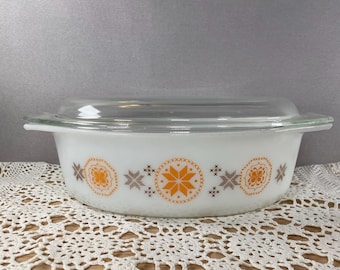 May include: White casserole dish with a clear glass lid. The dish features a decorative pattern of orange and brown starburst and floral designs. The dish has two handles and is sitting on a white lace tablecloth.