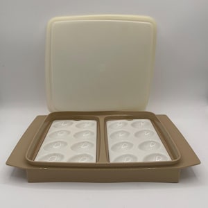 Puede incluir: Un recipiente de plástico beige para huevos con tapa a juego. El recipiente tiene dos bandejas blancas, cada una con hendiduras ovaladas para sujetar los huevos. La tapa es rectangular y se coloca encima del recipiente. El recipiente está diseñado para guardar huevos.