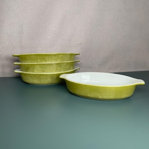 May include: Four oval-shaped, light green ceramic baking dishes with white interiors. The dishes are stacked in a group of three, with a fourth dish positioned separately. Each dish has small handles on either side.