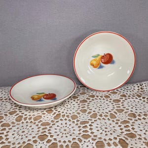 Könnte beinhalten: Zwei weiße Keramikschalen mit rotem Rand. Jede Schale hat ein gemaltes Design einer Birne und eines Apfels. Die Schalen stehen auf einer weißen Spitzendecke und einem grauen Hintergrund.