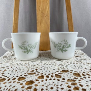 Può includere: Due tazze da caffè in ceramica bianca con un motivo floreale verde. Ogni tazza ha un manico arrotondato e una forma semplice ed elegante. Il motivo floreale comprende foglie e fiori. Le tazze sono esposte su una tovaglietta di pizzo bianco.