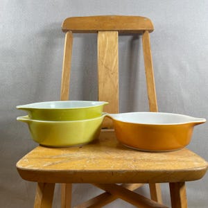 Op de afbeelding: Drie vintage Pyrex ovenschalen in tinten geel en oranje, met witte binnenkanten. De schalen zijn ovaal van vorm met handvatten aan elke kant. Ze zijn gestapeld op een houten stoel.