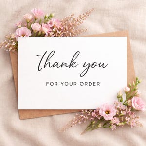 May include: A white card with the words "Thank you FOR YOUR ORDER" in black script is placed on a brown envelope. Pink flowers and greenery surround the card, set against a neutral fabric background.