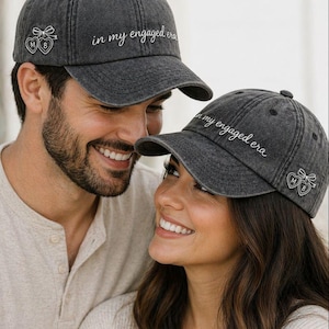 Puede incluir: Dos gorras de béisbol grises con el texto bordado "in my engaged era". Cada gorra presenta un diseño de corazón y lazo con las iniciales "M B". Las gorras son usadas por una pareja sonriente.