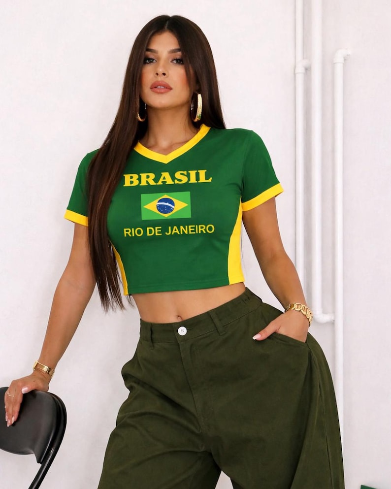 May include: A green crop top with yellow trim and the word "BRASIL" above a Brazilian flag graphic. The text "RIO DE JANEIRO" is below the flag. The top is paired with olive green pants.
