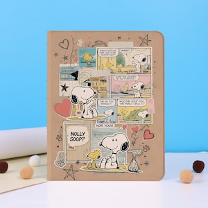 Op de afbeelding: Een bruine tablethoes met een collage van Snoopy en Woodstock strips. De hoes heeft hart-, ster- en pootafdrukstickers. Tekst bevat "NOLLY SOOP?" en stripballonnen.