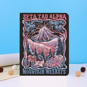 Puede incluir: Cuaderno negro con un diseño de paisaje de montaña colorido. La imagen presenta una cordillera con árboles, flores y el texto "ZETA TAU ALPHA MOUNTAIN WEEKET'S" en una fuente decorativa. El fondo es azul claro.