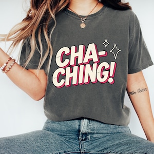 Könnte beinhalten: Ein anthrazitfarbenes Kurzarm-T-Shirt mit dem Schriftzug "CHA-CHING!" in einer fetten, cremefarbenen Schriftart mit rosa Umrandung und Schatteneffekt. Rechts neben dem Text auf der Brust befinden sich zwei kleine vierzackige Sternsymbole.