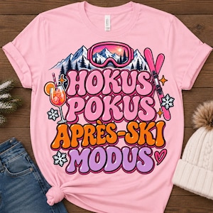 Könnte beinhalten: Ein hellrosa T-Shirt mit kurzen Ärmeln und einem Grafikdruck mit dem Text HOKUS POKUS APRÈS-SKI MODUS. Das Design zeigt ein Gebirge, eine rosa Skibrille, einen Cocktail mit einer Orangenscheibe, rosa Skier und weiße Schneeflocken.