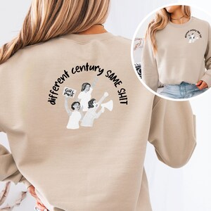 Könnte beinhalten: Beiges Sweatshirt mit einer Grafik auf dem Rücken, die drei protestierende Figuren mit den Worten "different century SAME SHIT" zeigt. Eine kleinere Grafik auf der rechten Brustseite vorne lautet "different century same shit."