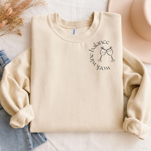 Könnte beinhalten: Cremefarbenes Sweatshirt mit dem Schriftzug "work wine balance" in einem kreisförmigen Design, begleitet von zwei Weingläsern. Das langärmlige Oberteil ist gefaltet, und ein Hut und Jeans sind im Hintergrund zu sehen.