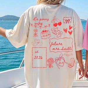 Könnte beinhalten: Cremefarbenes T-Shirt mit roten grafischen Illustrationen und Text. Das Design enthält Sätze wie "it's giving wifey" und "future mrs. Smith" sowie Zeichnungen von Herzen, einem Kuchen und einem Blumenstrauß. Das Jahr "2026" ist ebenfalls abgebildet.