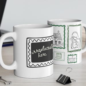 May include: Two white ceramic mugs. One mug has the text "irreplaceable here" in a black chalkboard-style frame. The other mug features green-framed sketches of a house, a person, and a calendar. The mugs are on a light-colored surface.