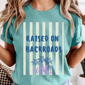 May include: Teal t-shirt with the text "RAISED ON BACKROADS" in blue, framed by vertical white stripes. Below the text are blue and purple floral illustrations. The shirt has short sleeves and a crew neck.