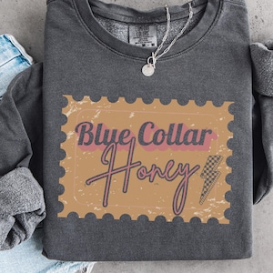 May include: Dark gray sweatshirt with the text "Blue Collar Honey" in a stamp-like design. The design includes a lightning bolt graphic. A silver necklace with a pendant is visible. Rolled-up denim jeans are on the left.