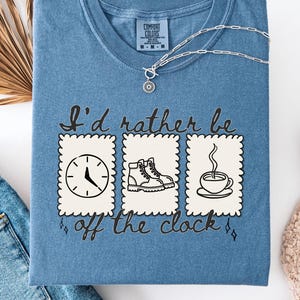 May include: A blue Comfort Colors t-shirt with the phrase "I'd rather be off the clock" and illustrations of a clock, hiking boot, and coffee cup. A silver necklace and jeans are also visible.
