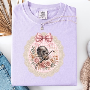 May include: A light purple Comfort Colors t-shirt featuring a decorative design. The design includes a welding helmet, a pink bow, and floral elements. A silver necklace and white shoes are also visible.