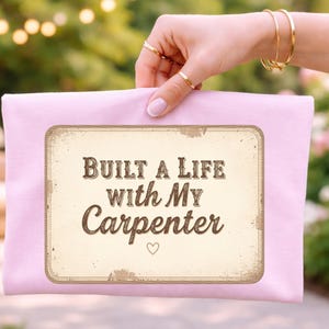 May include: A light pink clutch with a rectangular design that reads "Built a Life with My Carpenter" in brown text. The design has a vintage, distressed look, with a small heart symbol. The background is blurred.