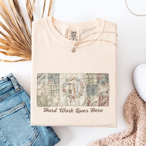 Puede incluir: Camiseta color crema con un diseño de patchwork con herramientas de jardinería y el texto "Hard Work Lives Here". El diseño incluye un martillo, una paleta y otras herramientas, rodeadas de motivos florales.
