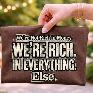 May include: A brown clutch with the text "We're Not Rich in Money. WE'RE RICH. IN EVERYTHING. Else." in a bold, distressed font. The clutch is held by a hand with gold rings and bracelets, set against a blurred outdoor background.