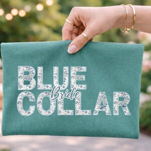May include: A teal-colored clutch with the words "BLUE Bride COLLAR" in white, floral-filled letters. The clutch is held up by a hand with gold rings and bracelets, with a blurred outdoor background.