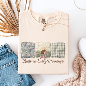 May include: Cream-colored t-shirt with a patchwork design featuring floral, sunrise, and plaid patterns. The text "Built on Early Mornings" is printed below the patches. A gold necklace and a pair of blue jeans are also visible.