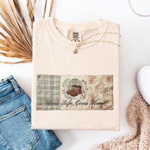 May include: Cream-colored t-shirt with a decorative patch featuring a plaid pattern, a jacket and boots illustration, and a floral design. The text "Work Safe, Come Home" is below the illustrations.